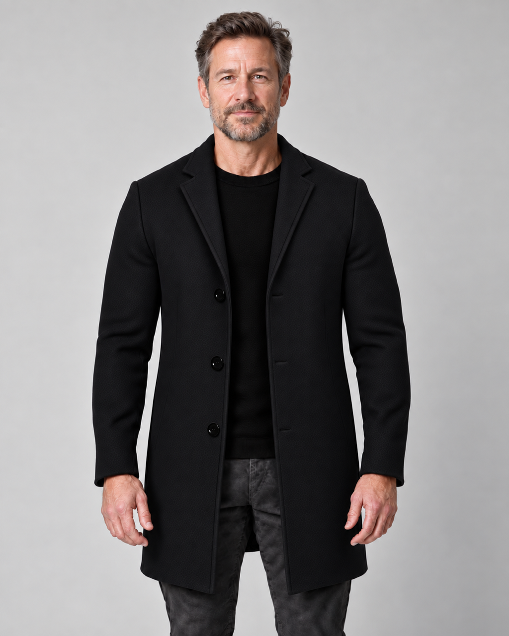 Homme portant un manteau noir à trois boutons, vu de face sur un fond gris uni.