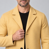 Gros plan de face d’un homme portant un manteau jaune moutarde à revers et boutons noirs.